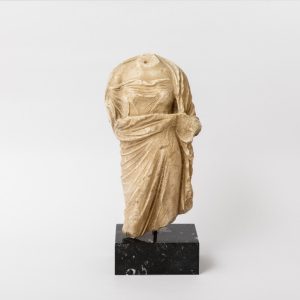 Αναπαράσταση γυναικείου σώματος με ιμάτιο το οποίο συγκρατείται από το ένα χέρι της και είναι ελαφρώς σηκωμένο.