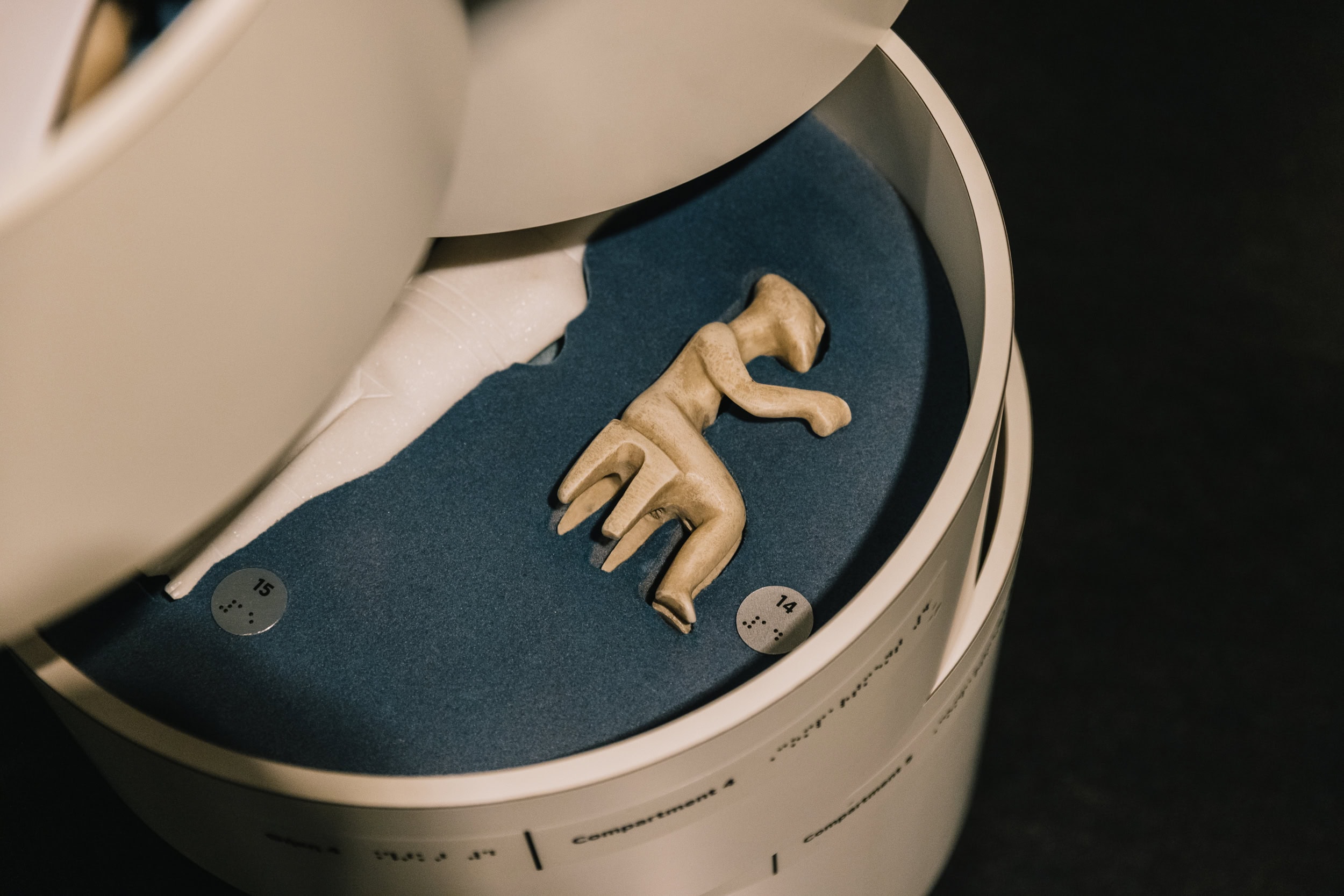 Image of the mobile tactile display case The image shows a small cycladic figurine displayed inside a museum case. The object appears to be made of clay or stone and has an abstract, stylized form resembling a human figure in a dynamic pose. The figurine rests on a dark blue surface inside a cylindrical display.
