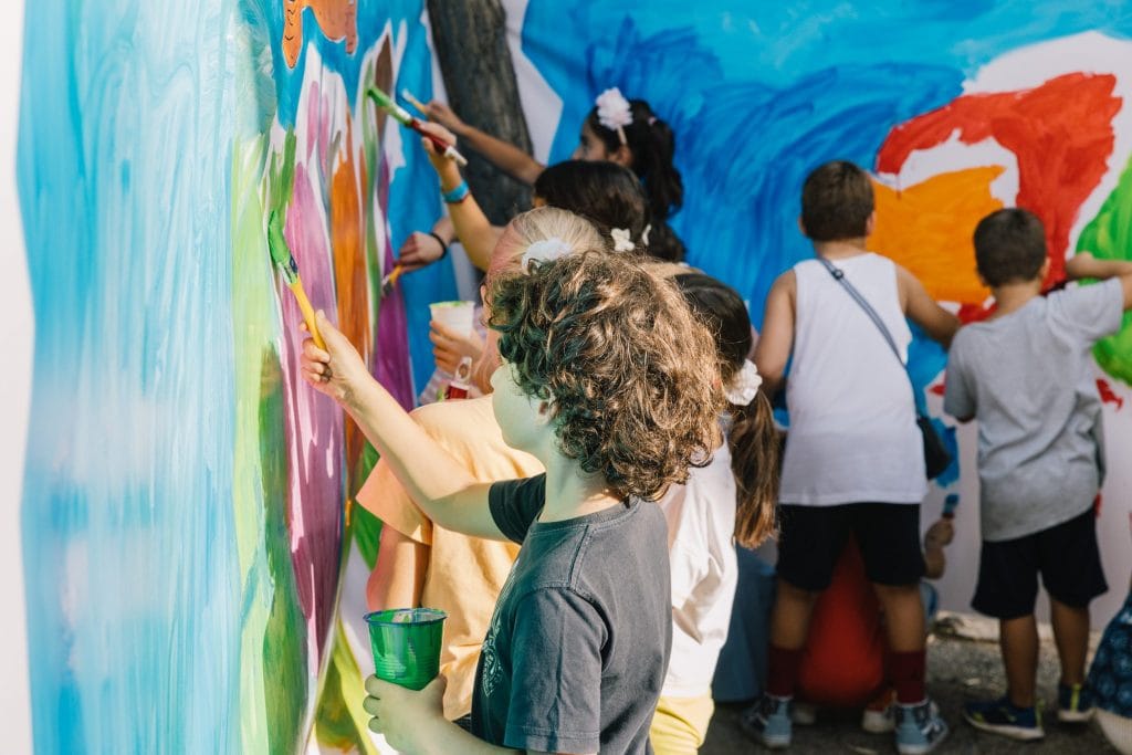Boys and girls stand in front of two large walls, painting them with brushes and paints.